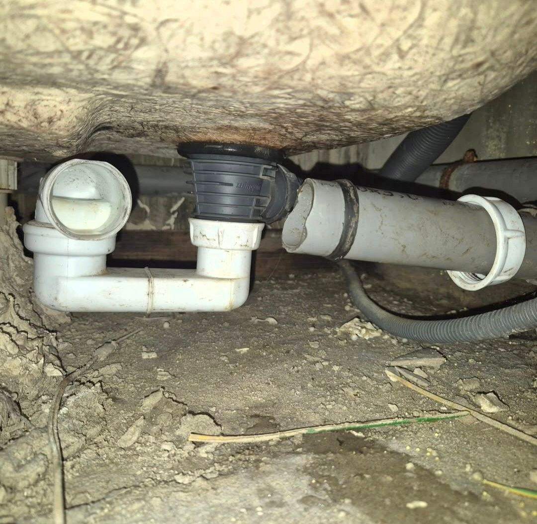 Sous une baignoire, un système d'évacuation en plastique est déconnecté d'un tuyau, laissant apparaître de la saleté sur le sol.