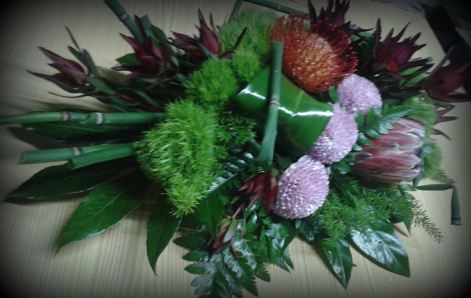 Arranjo floral com flores verdes, rosas e laranjas, bambu e folhagem sobre uma superfície de madeira.