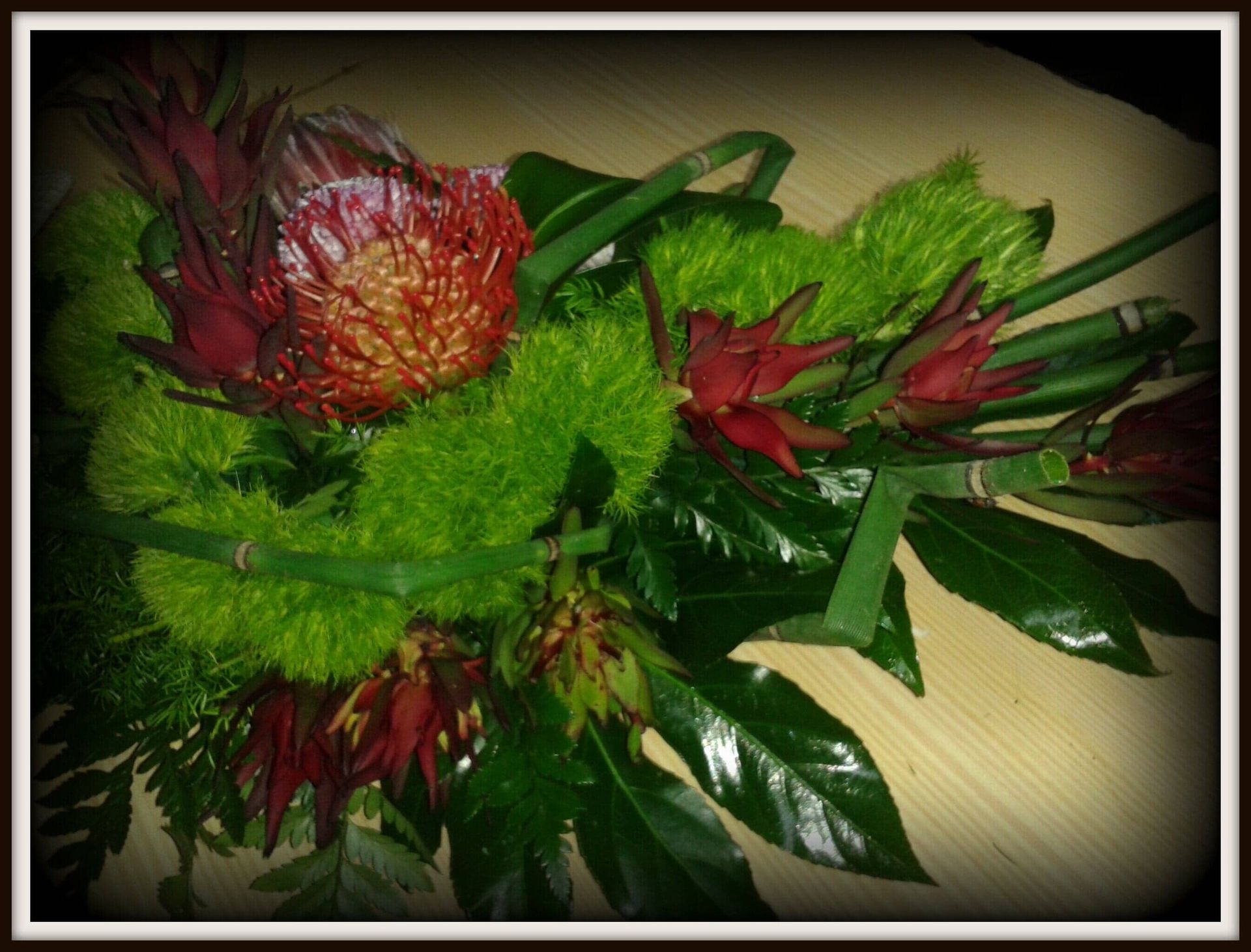 Detalhe de um arranjo floral: flores vermelhas e verdes, folhagem verde exuberante, sobre uma superfície marrom clara.