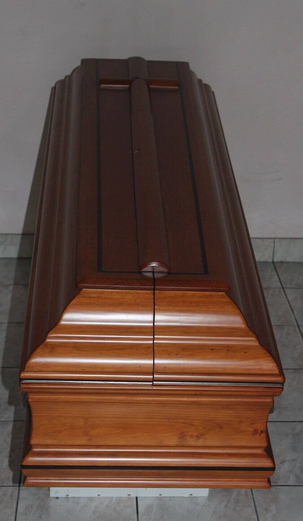 Um caixão de madeira fechado, em tons de marrom e claro, com uma alça de metal, dentro de um quarto.