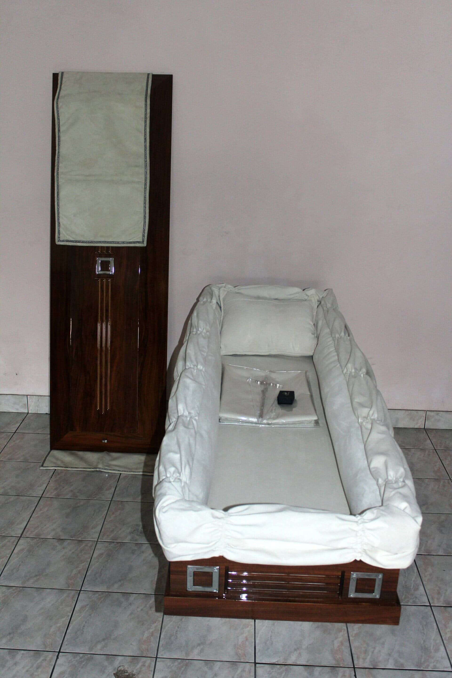 Caixão aberto com interior branco, tampa apoiada ao lado, sobre piso de azulejos.