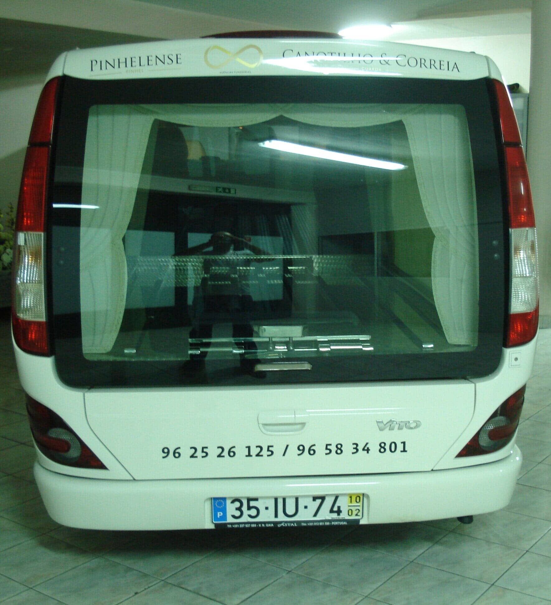 Vista traseira de um carro funerário branco com grandes janelas. Possui cortinas internas.