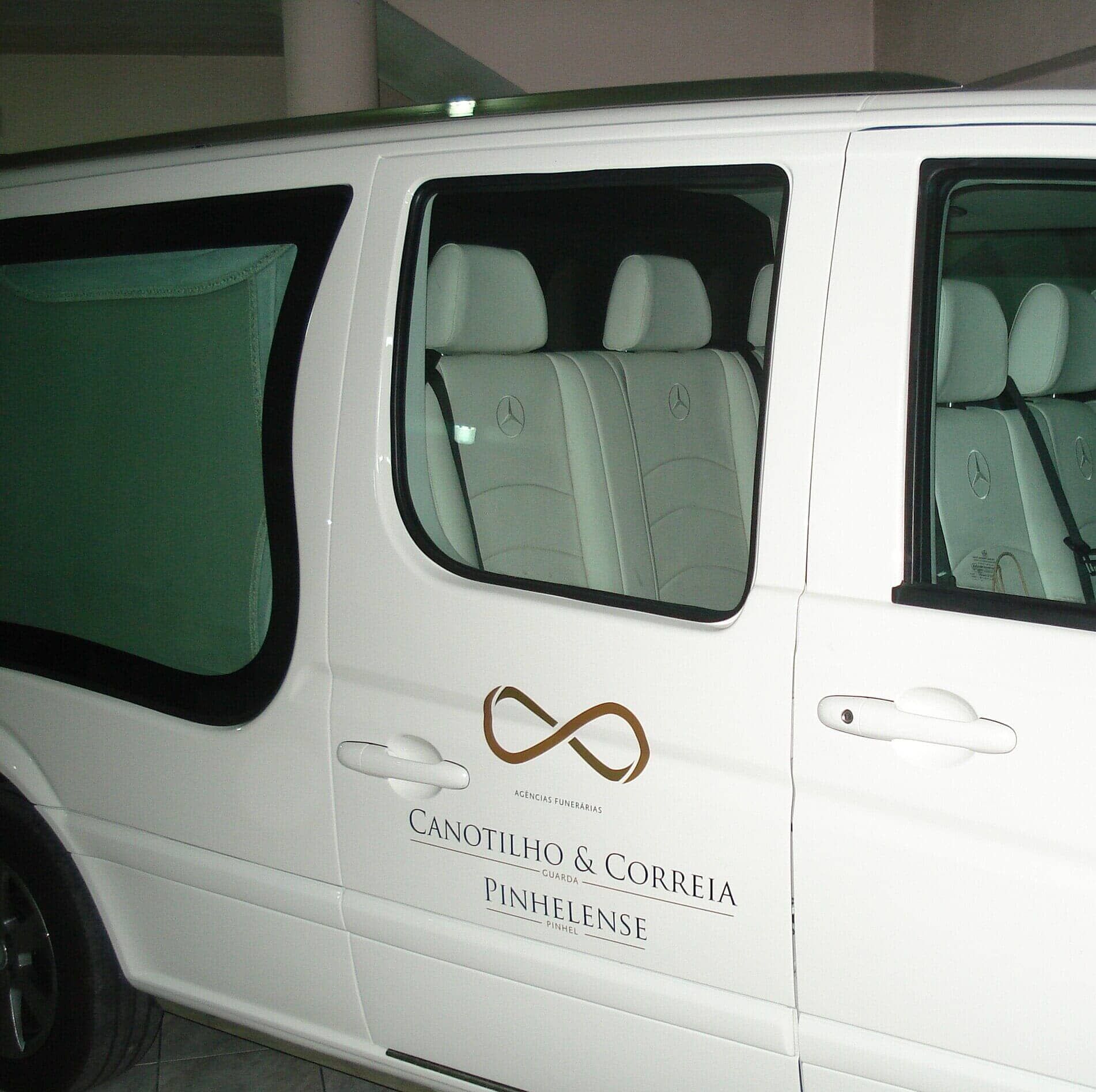 Carro funerário branco com o logotipo do infinito, estacionado ao ar livre.