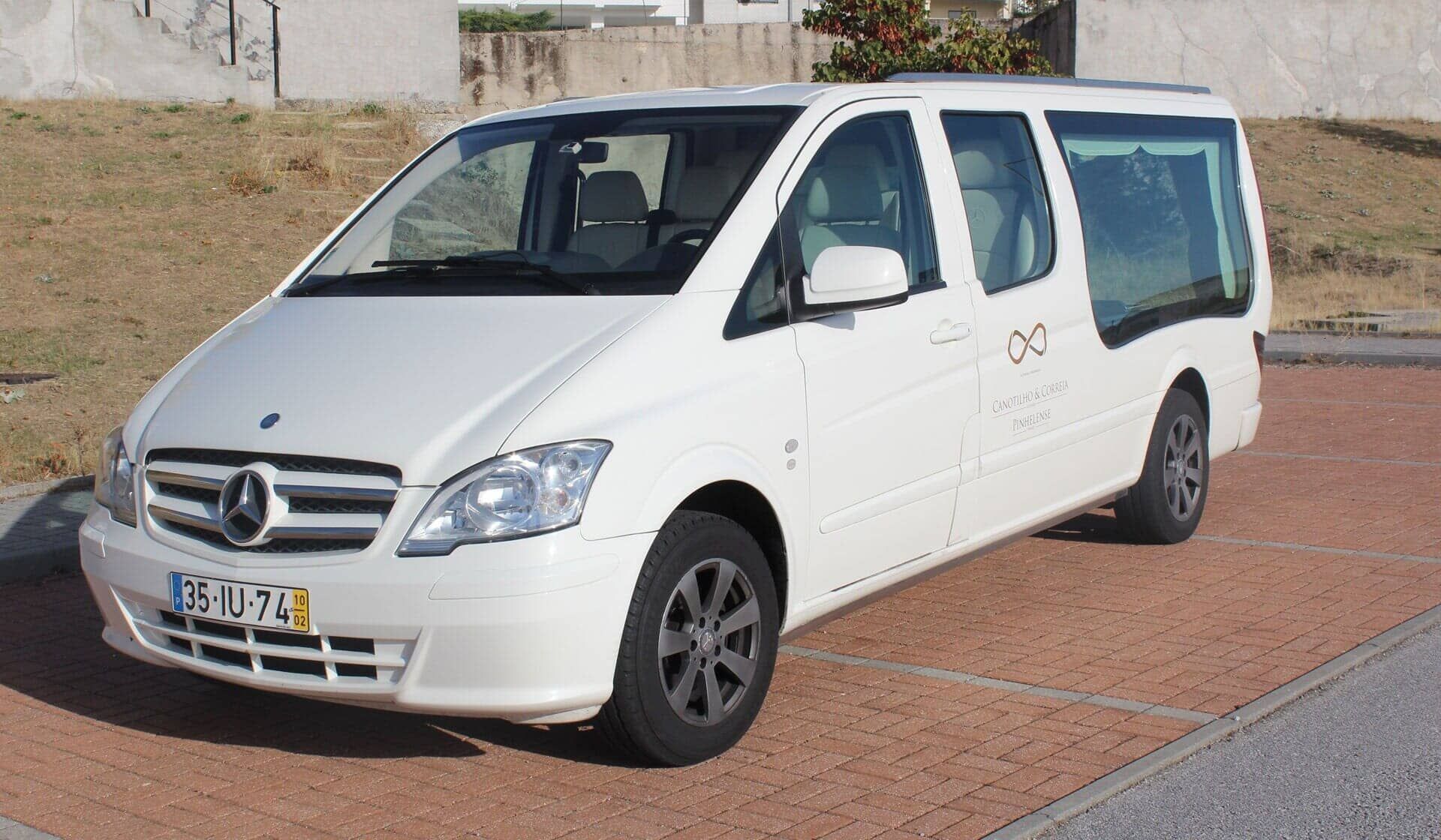 Carro funerário Mercedes branco estacionado sobre calçada de tijolos.
