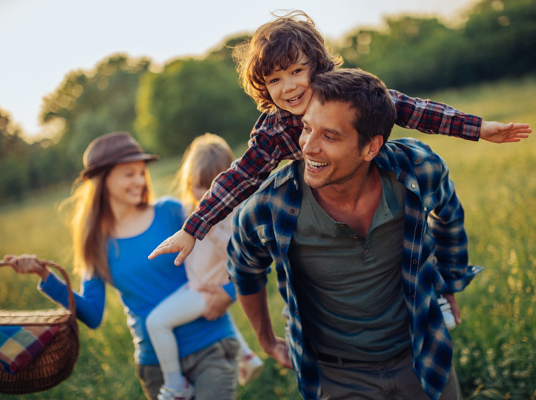 Familia en un picnic en un campo soleado: papá cargando a un niño, mamá cargando a otro niño, sonriendo.