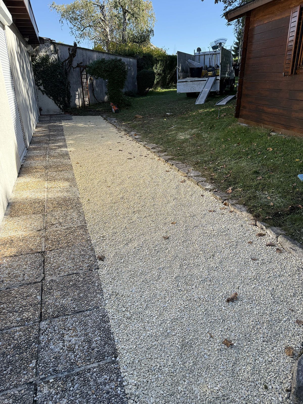 Chemin de gravier à côté d'un mur et d'une pelouse, menant vers une structure en bois et un camion sous un ciel bleu.