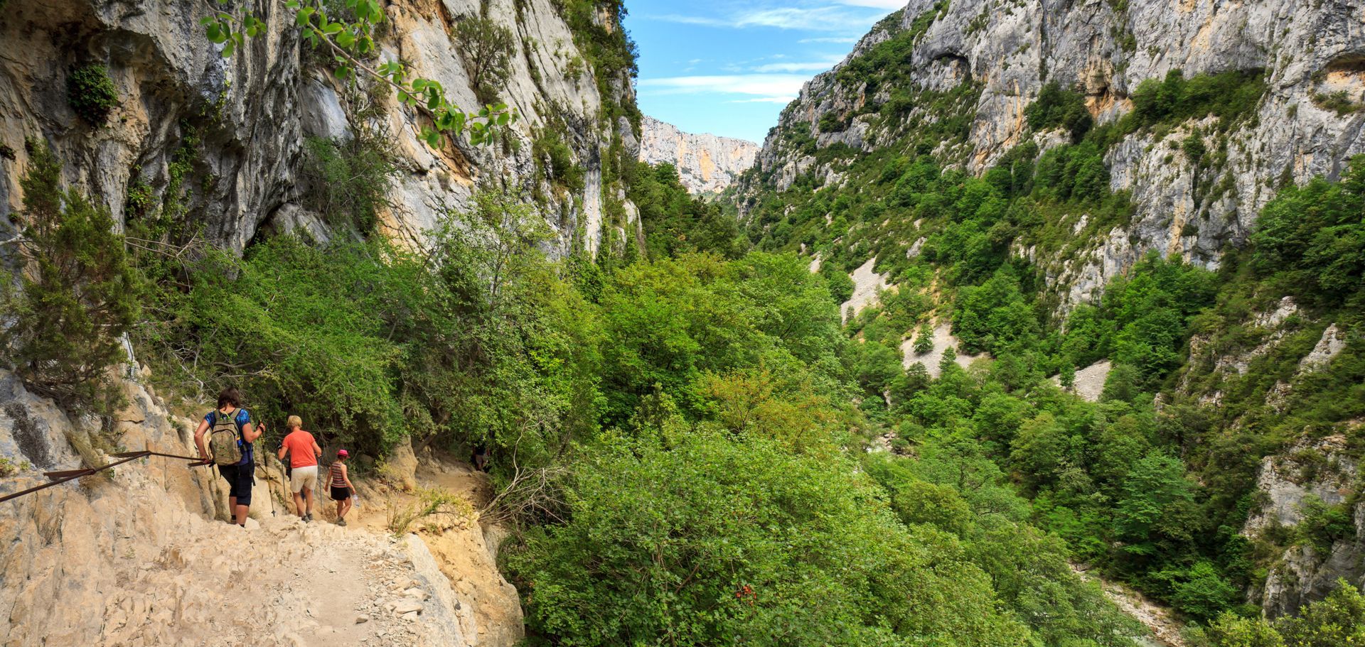 Randonneurs sur un sentier étroit à flanc de falaise, dans un canyon verdoyant, entouré de montagnes rocheuses.
