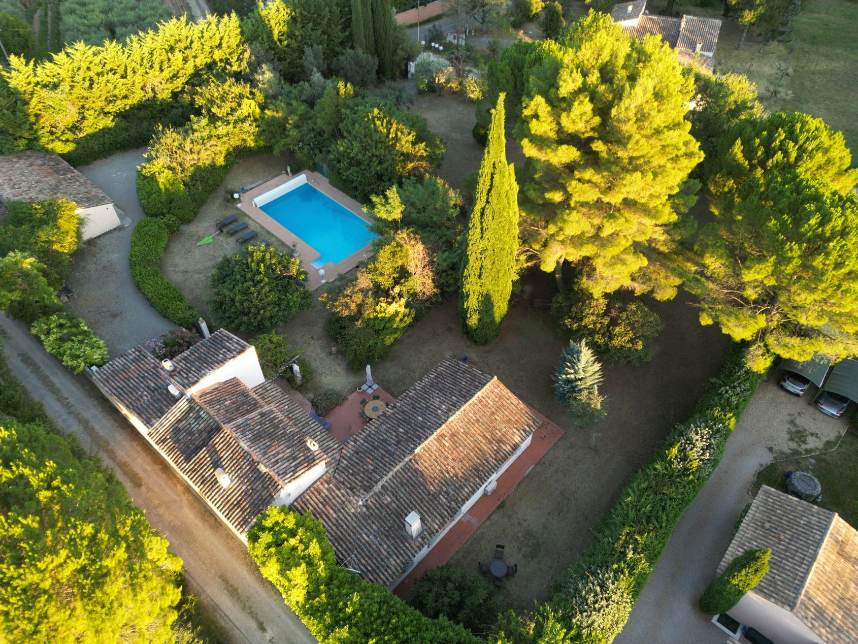 Vue aérienne d'une maison, d'une piscine et d'un jardin entourés de grands arbres baignés d'une douce lumière du soleil.