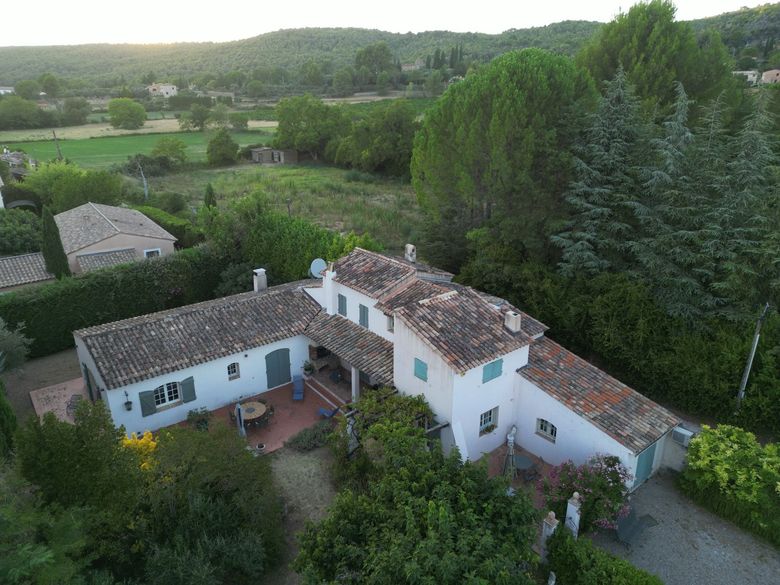 Vue aérienne d'une maison au toit de tuiles, entourée d'arbres denses et de champs verdoyants.