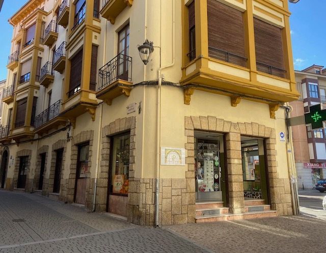 Edificio de esquina con farmacia en la planta baja; paredes color canela, contraventanas marrones y detalles en piedra.