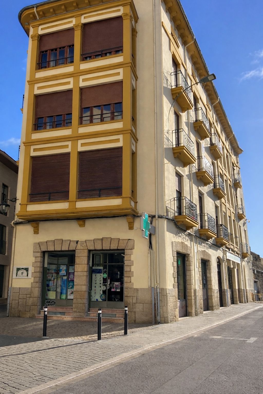 Edificio esquinero con tienda, molduras amarillas, persianas marrones y balcones de hierro forjado. Acera de piedra, cielo azul.