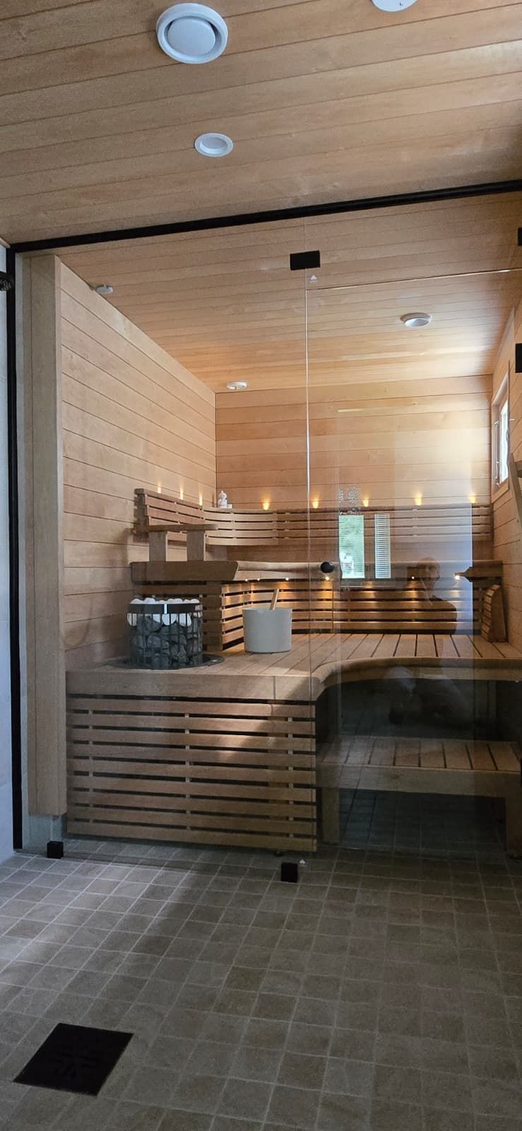 Puupaneloitu sauna, jossa on lauteet, kiuas ja laattalattia. Sauna näkyy lasioven läpi