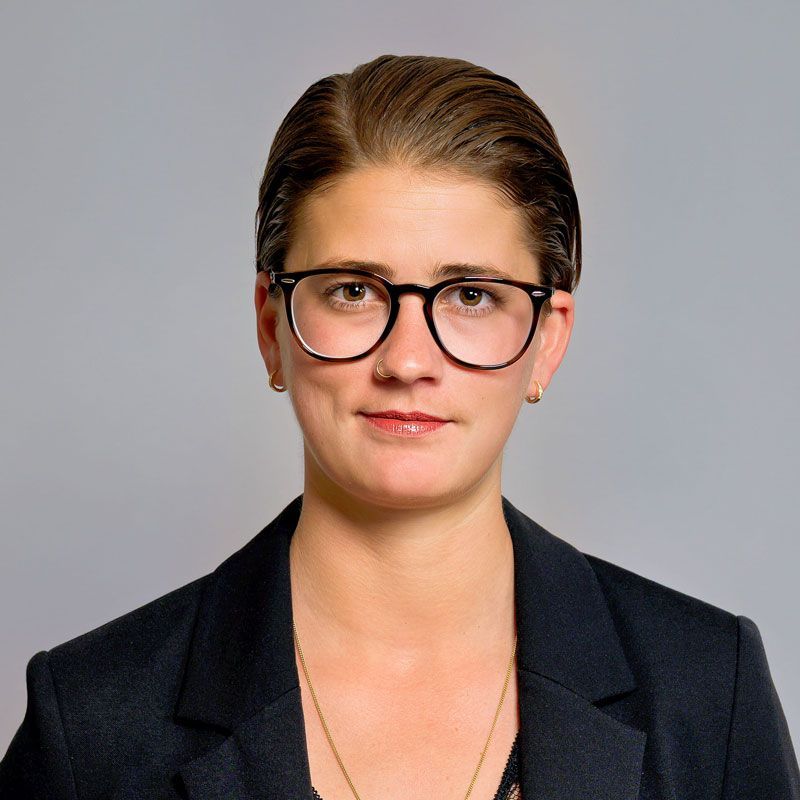 Eine Person mit Brille und dunkler Weste steht in einem Lagerhallengang mit hohen Regalen.