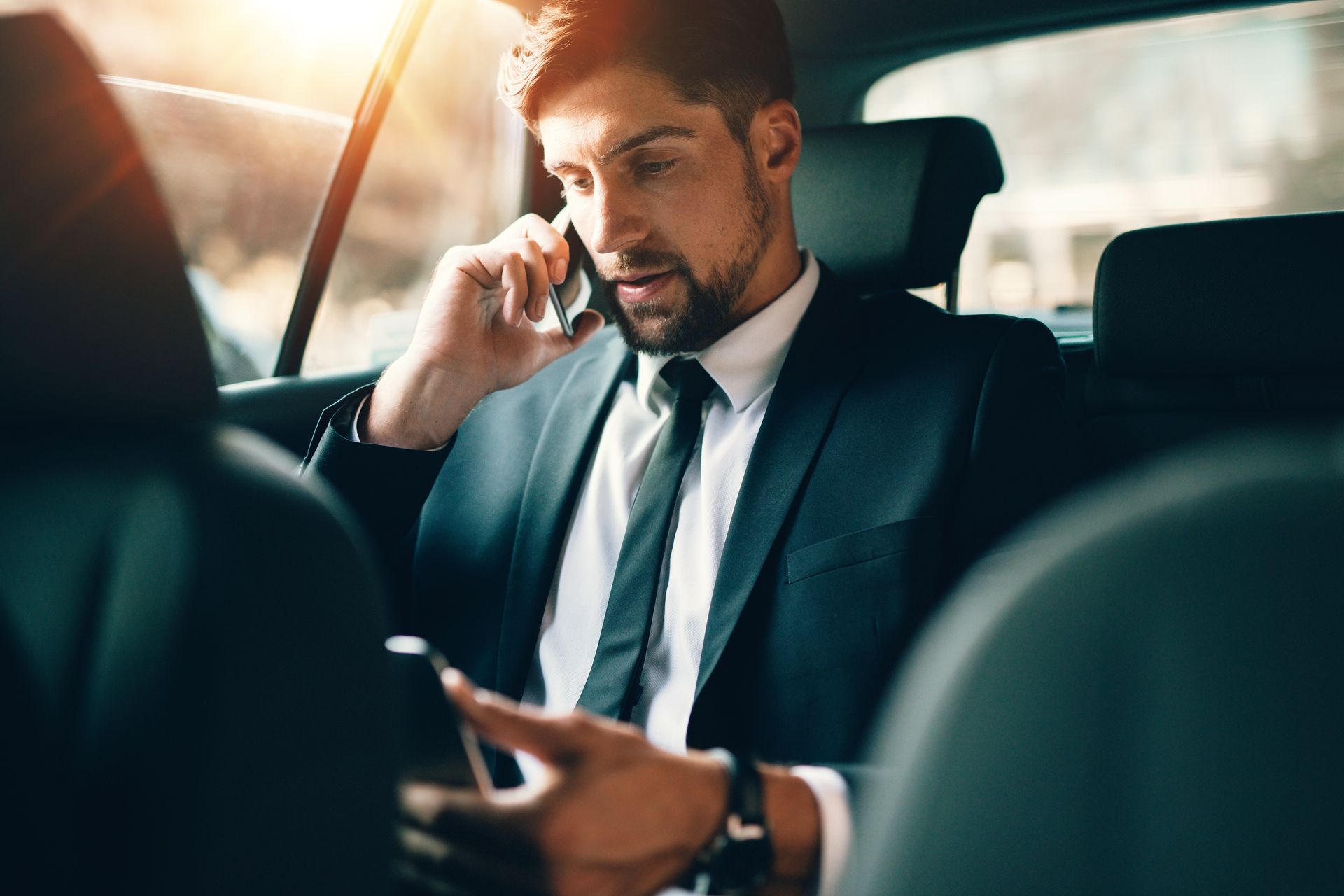 Un homme en costume sombre parle au téléphone dans une voiture, les yeux rivés sur son smartphone.