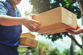 Un livreur remet un colis en carton à une autre personne à l'extérieur, avec d'autres cartons en arrière-plan.
