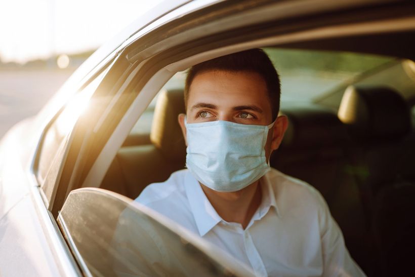 Un passager avec un masque, assis dans sa voiture, regarde par la fenêtre la douce lumière du soleil.