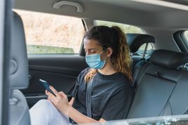 Une personne assise à l'arrière d'une voiture porte un masque bleu et regarde son téléphone.