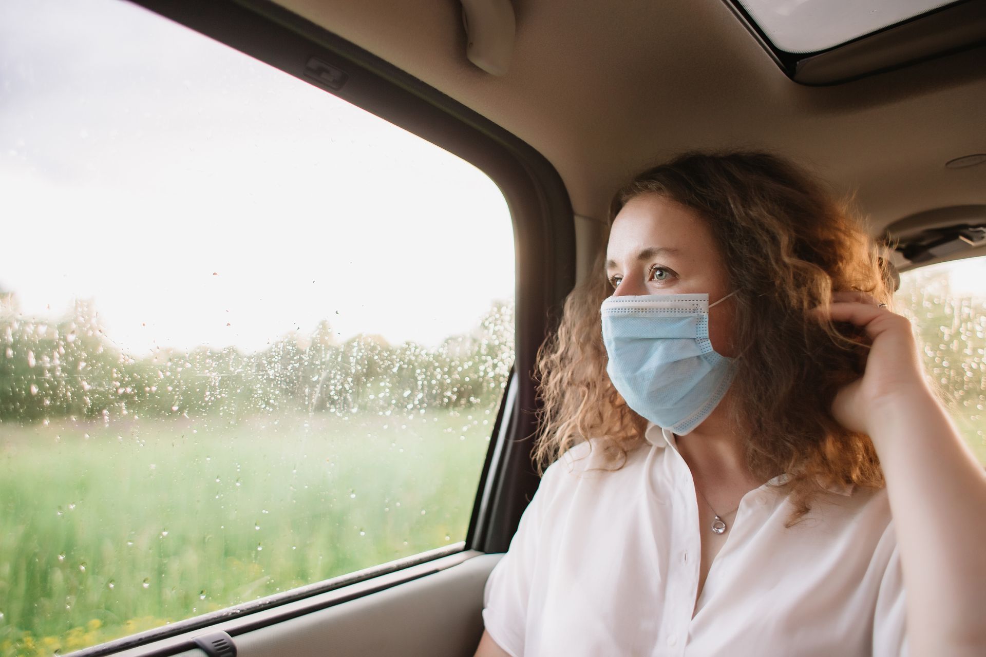 Une personne masquée assise près de la fenêtre d'une voiture, regardant un paysage pluvieux.