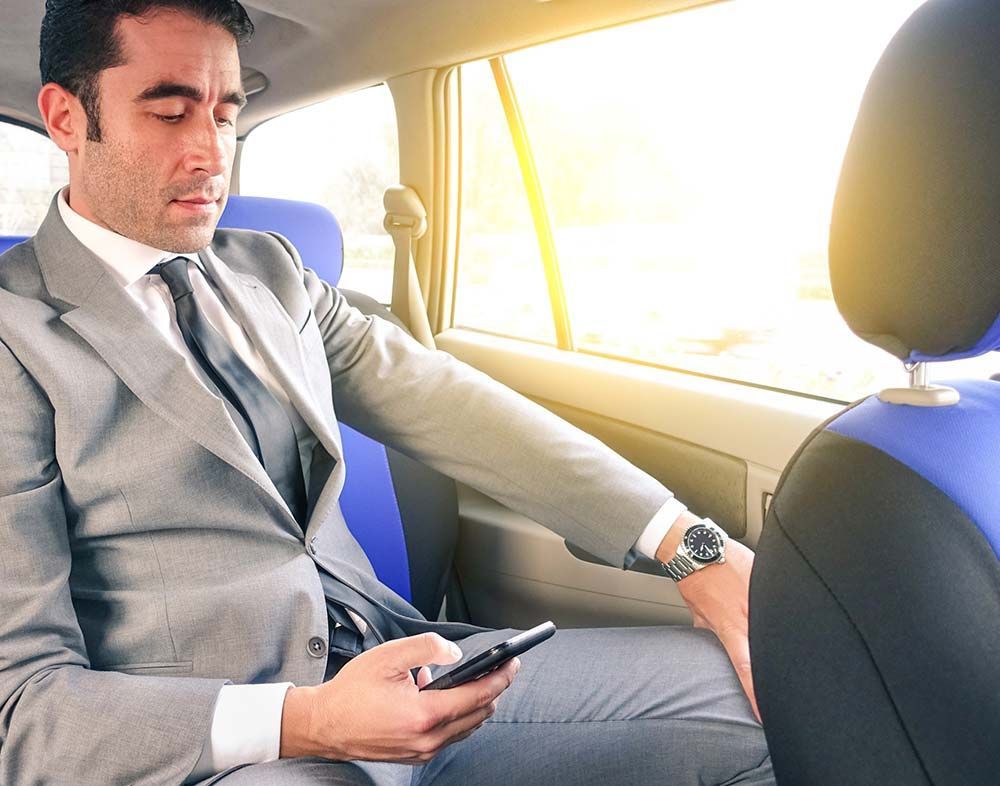 Un hombre con traje y corbata está sentado en el asiento trasero de un automóvil mirando su teléfono celular.