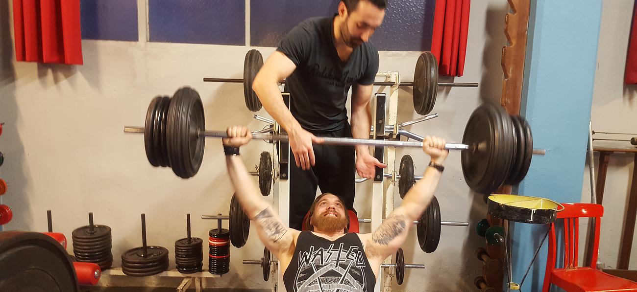 Homme avec barbe et tatouages allongé sur le dos en train de soulever des haltères aidé par un coach debout derrière