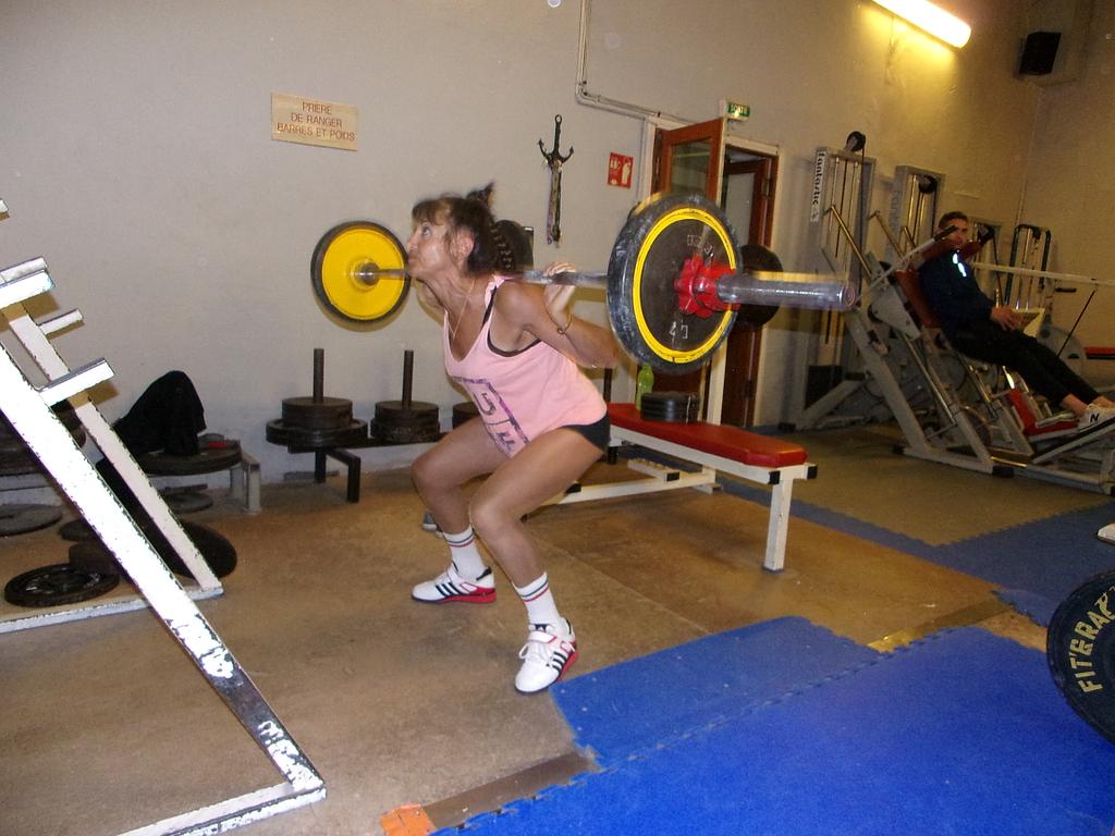 Femme debout en train de soulever sur son dos de lourdes haltères entourée de machines de musculation