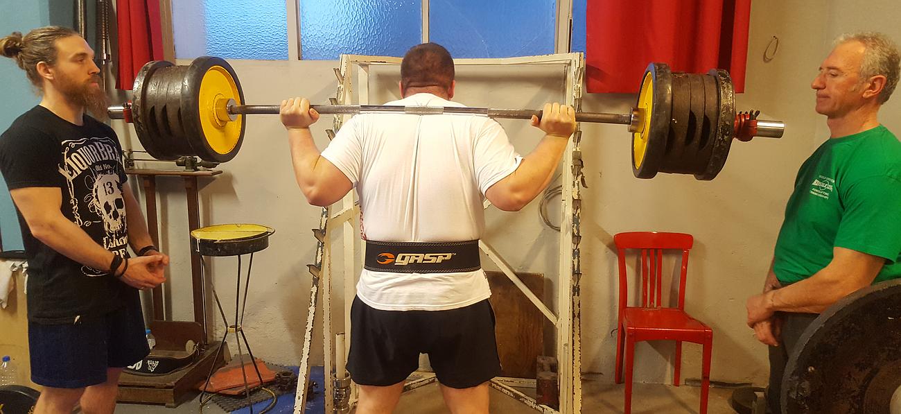 Homme de dos debout soulevant plusieurs poids sur une haltère, encadré par deux coachs