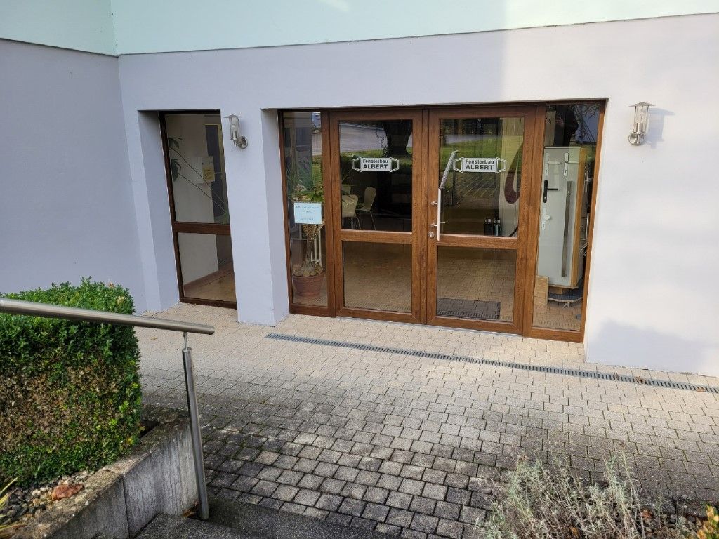 Außenansicht des Gebäudes mit einer Holztür am Eingang und zwei Seitentüren. Kopfsteinpflasterweg und Grünflächen davor.