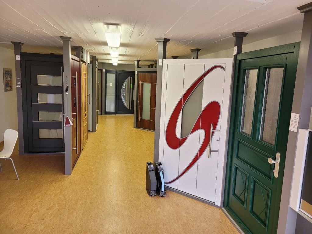 Innenraum eines Ausstellungsraums mit verschiedenen ausgestellten Türen. Holzboden, weiße Decke mit Lichtern und Säulen.