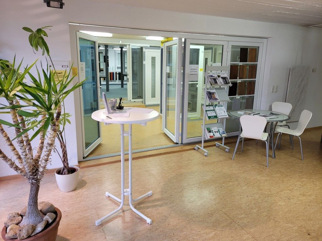 Innenausstellungsraum mit Türen. Weißer runder Tisch, Stühle, Topfpflanze und Ausstellungsregal.