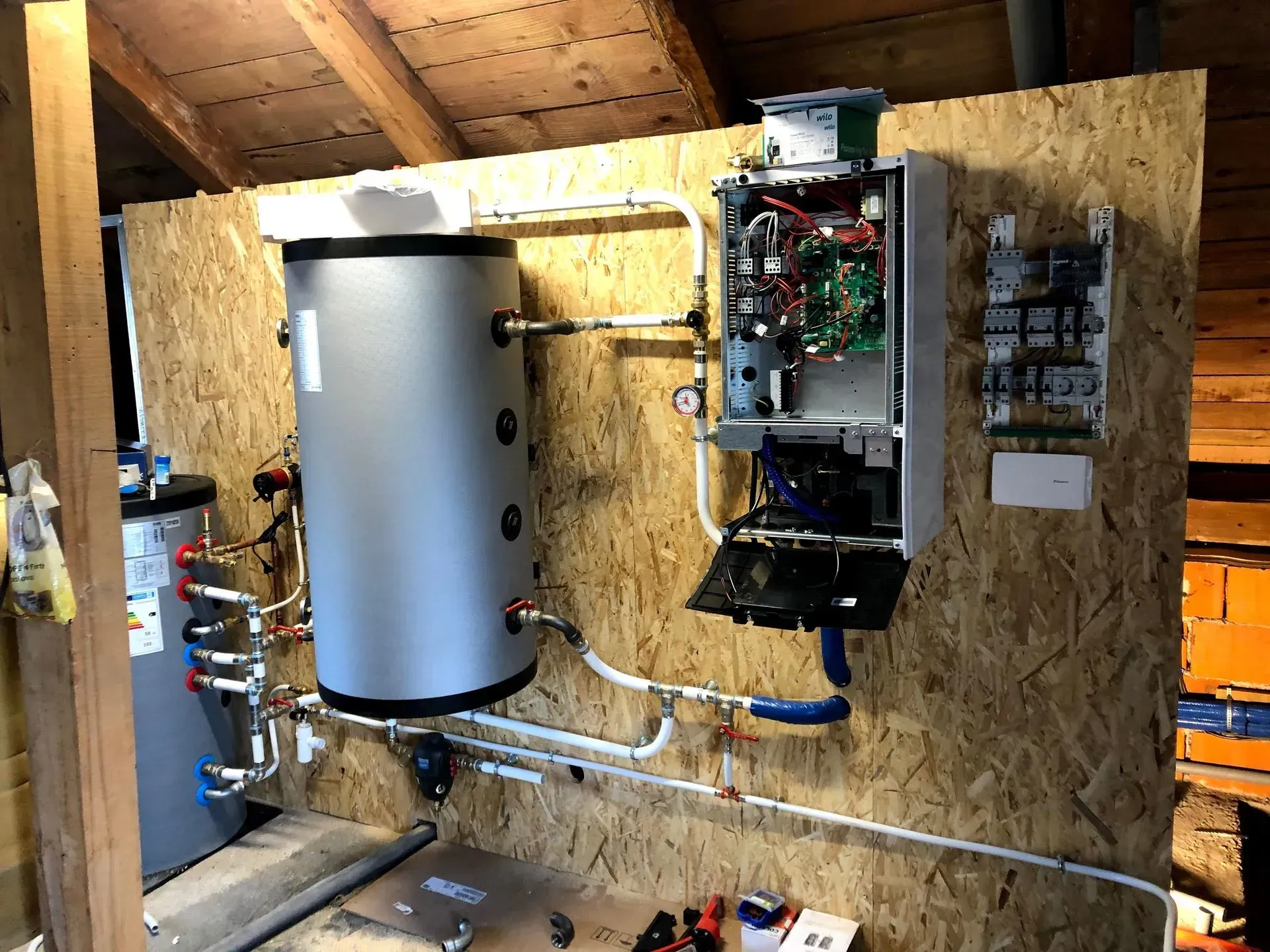 Chauffe-eau et chaudière fixés sur des panneaux de bois dans un grenier. Câblage, tuyaux et réservoirs apparents.