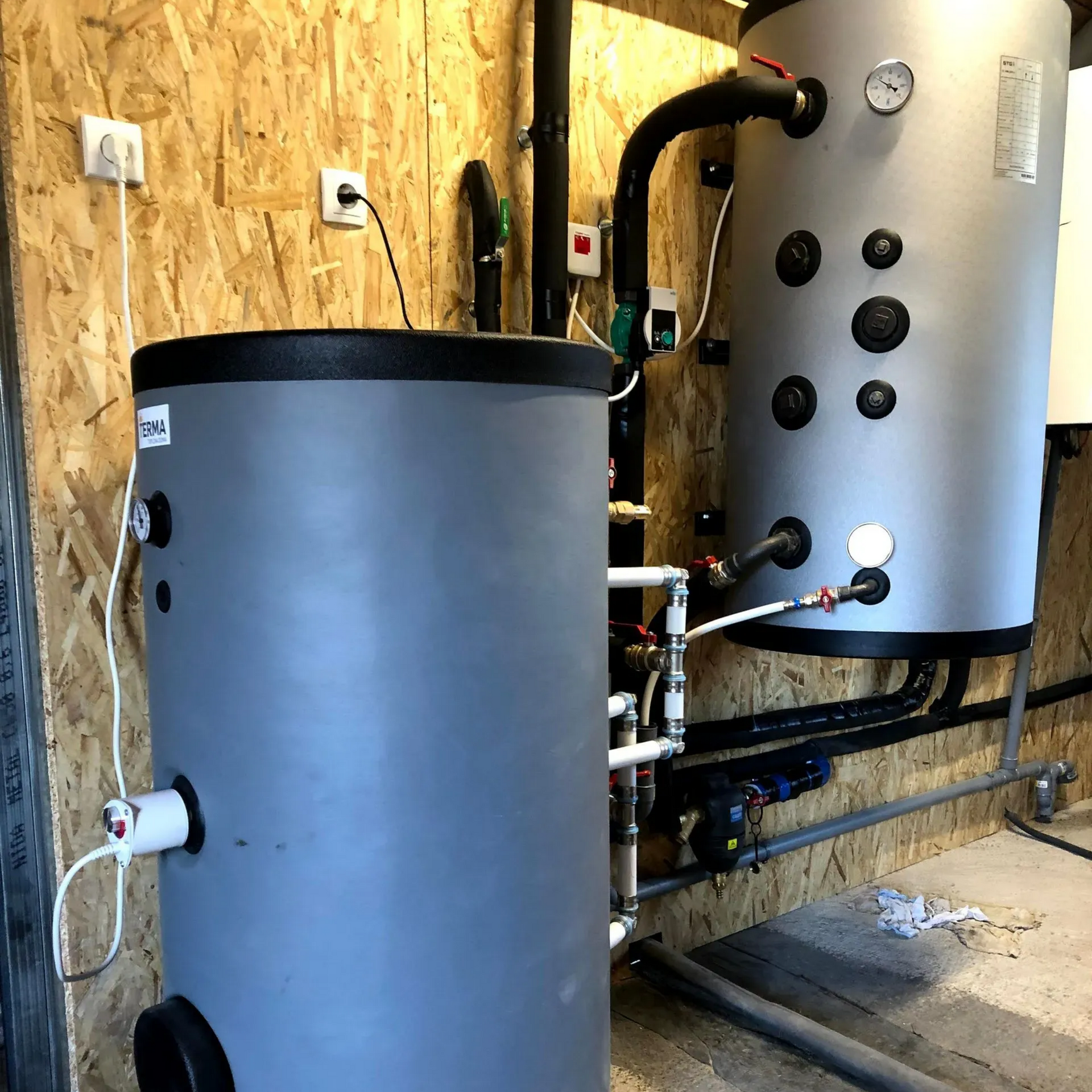 Deux grands réservoirs d'eau gris avec des tuyaux et des vannes noirs dans une buanderie aux murs en contreplaqué.