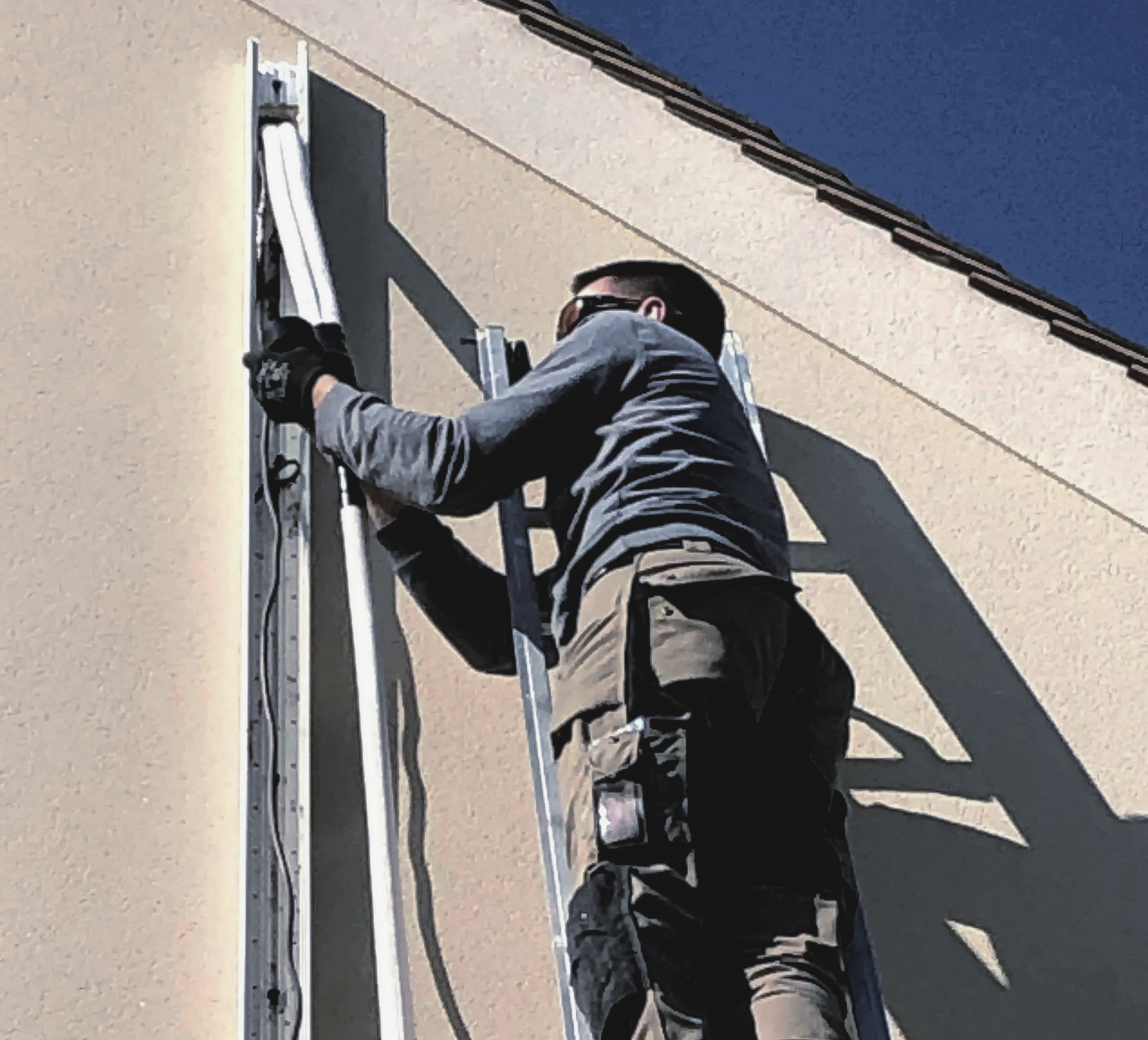 Une personne sur une échelle installe une gouttière blanche sur la façade beige d'un bâtiment.