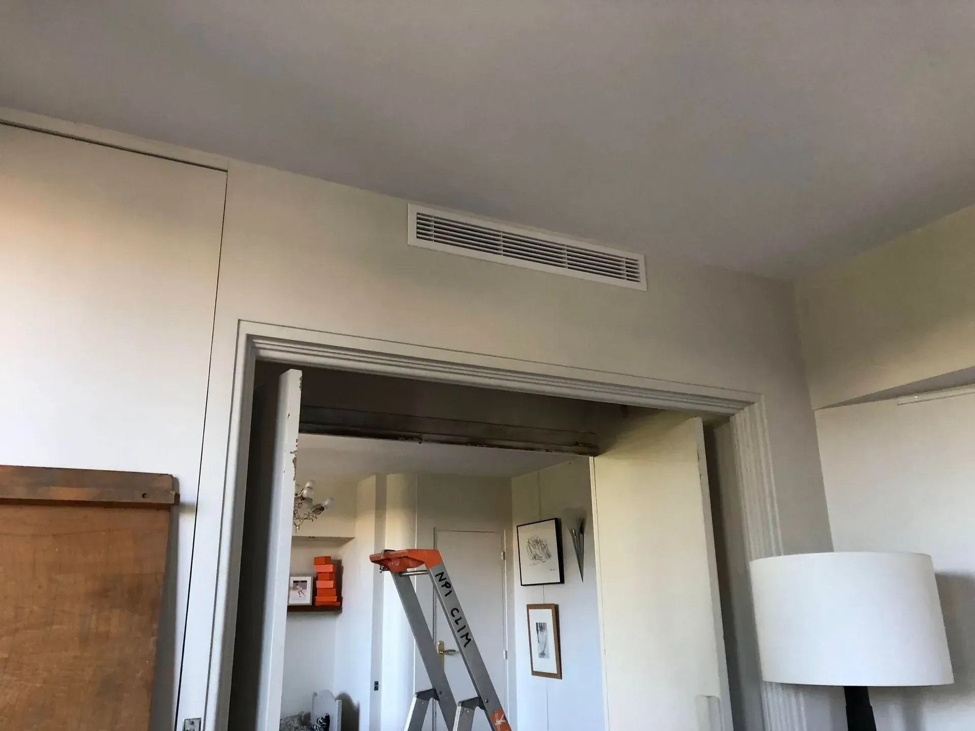 Grille d'aération au plafond au-dessus d'une porte ouverte ; un escabeau est visible dans la pièce adjacente.