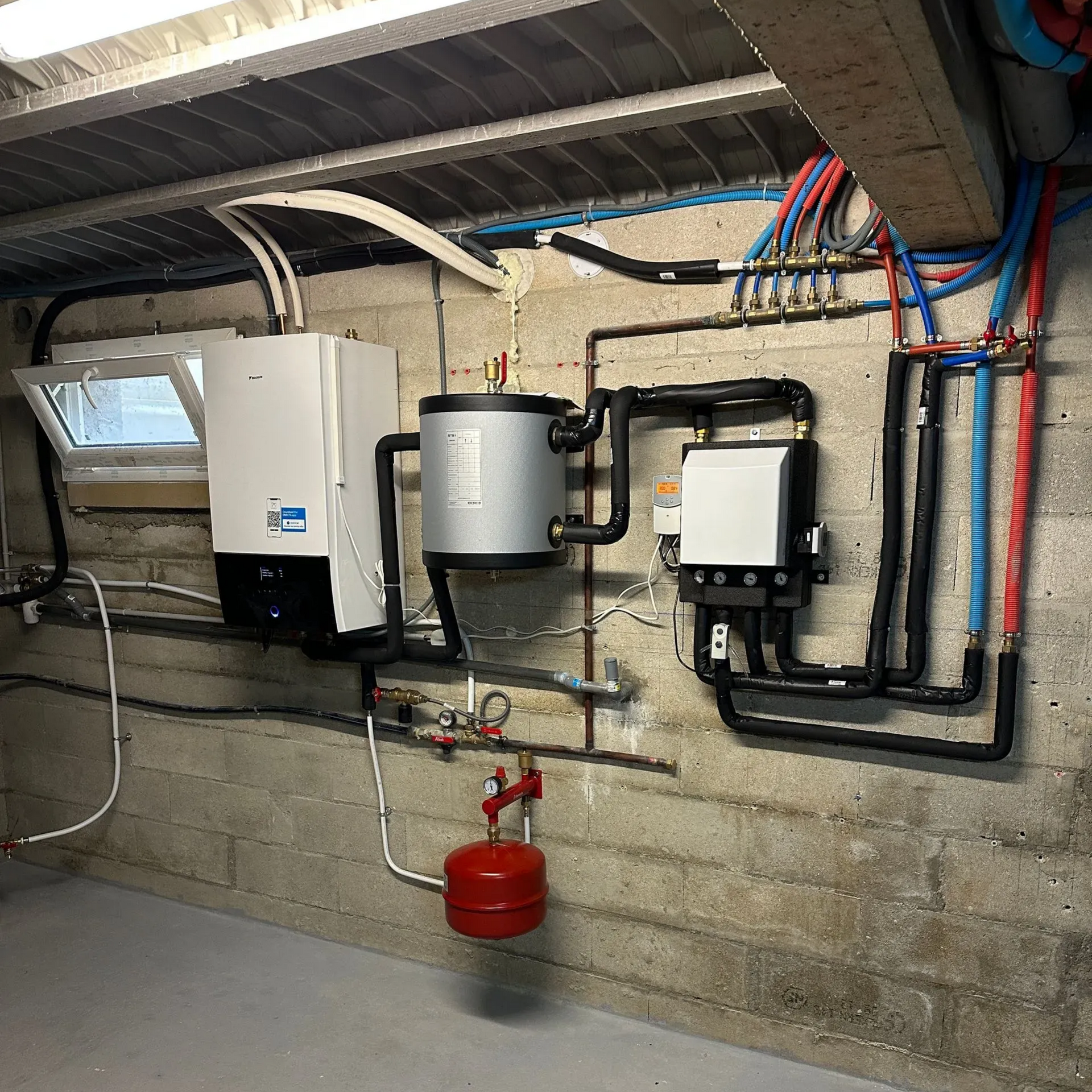 Équipements du système de chauffage sur un mur en béton : chaudière, chauffe-eau, vase d’expansion rouge et tuyaux isolés.