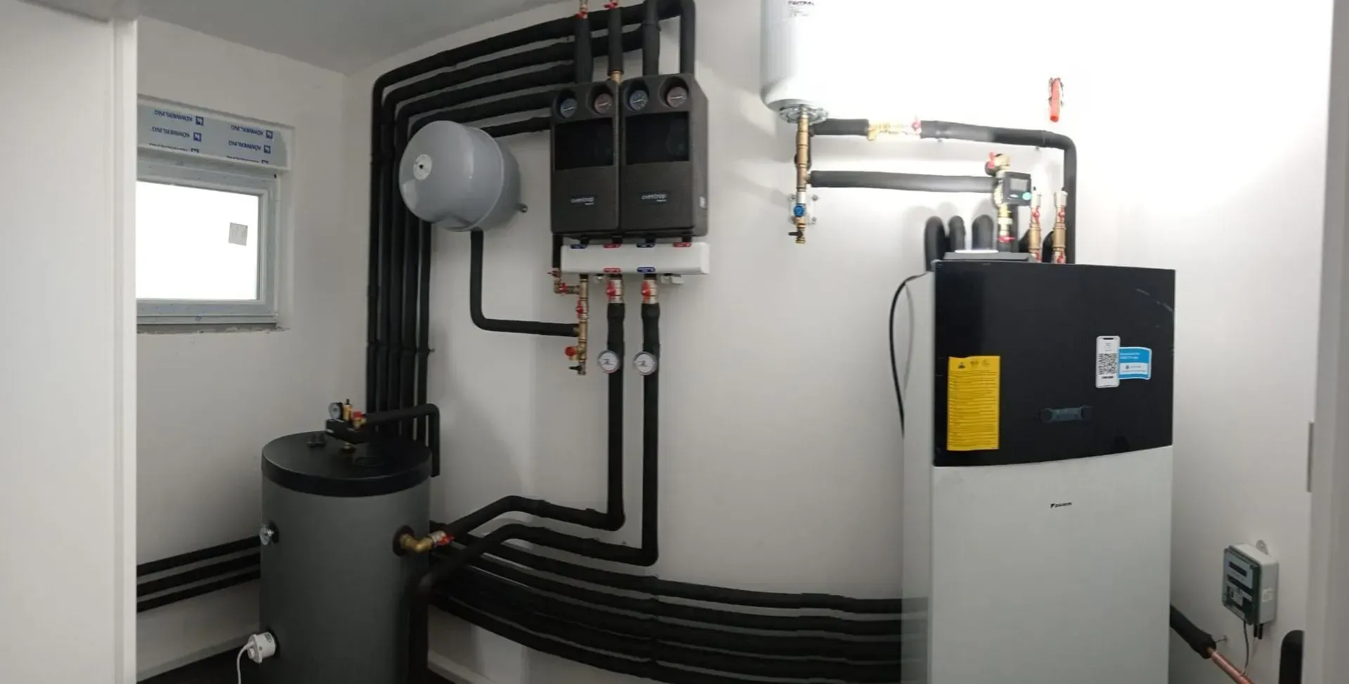 Local technique du système de chauffage avec tuyaux noirs, chauffe-eau gris et pompe à chaleur contre des murs blancs.