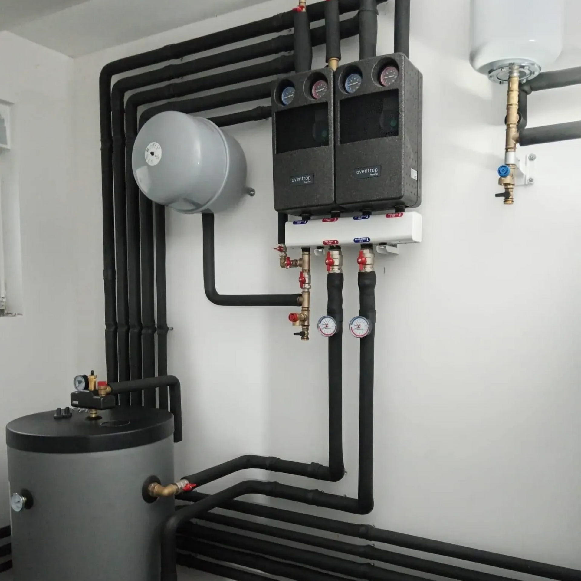 Système de chauffage avec tuyaux isolés noirs et divers réservoirs et manomètres montés sur un mur blanc.