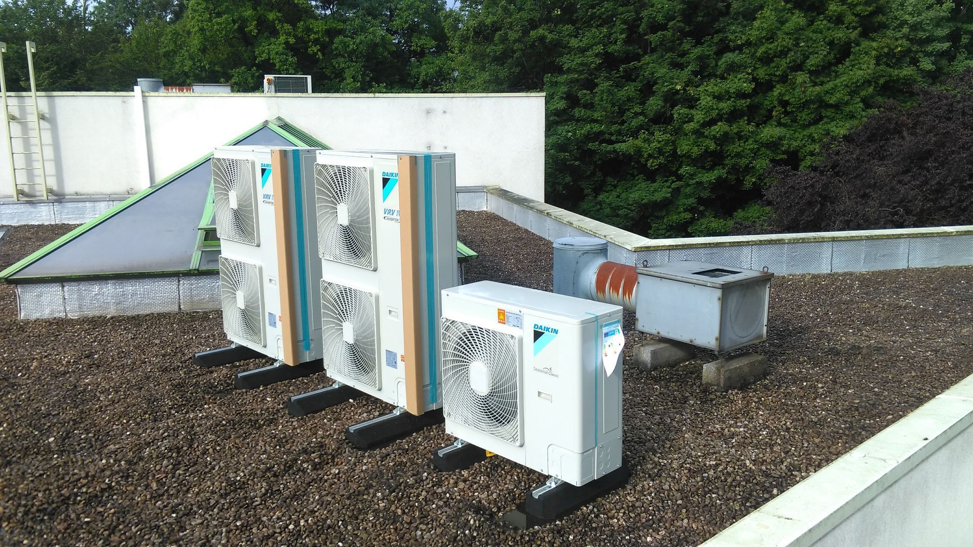 Vue sur un toit-terrasse avec plusieurs unités de climatisation installées sur une surface gravillonnée. Des arbres verts et un bâtiment blanc se dressent en arrière-plan.