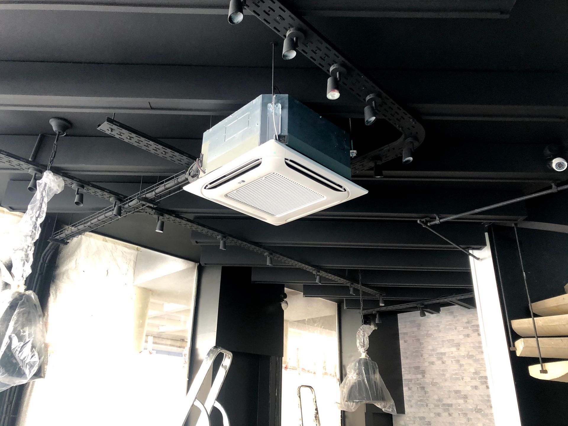 Unité de climatisation fixée au plafond dans une pièce avec poutres noires, éclairage sur rail et fenêtres sans rideaux.