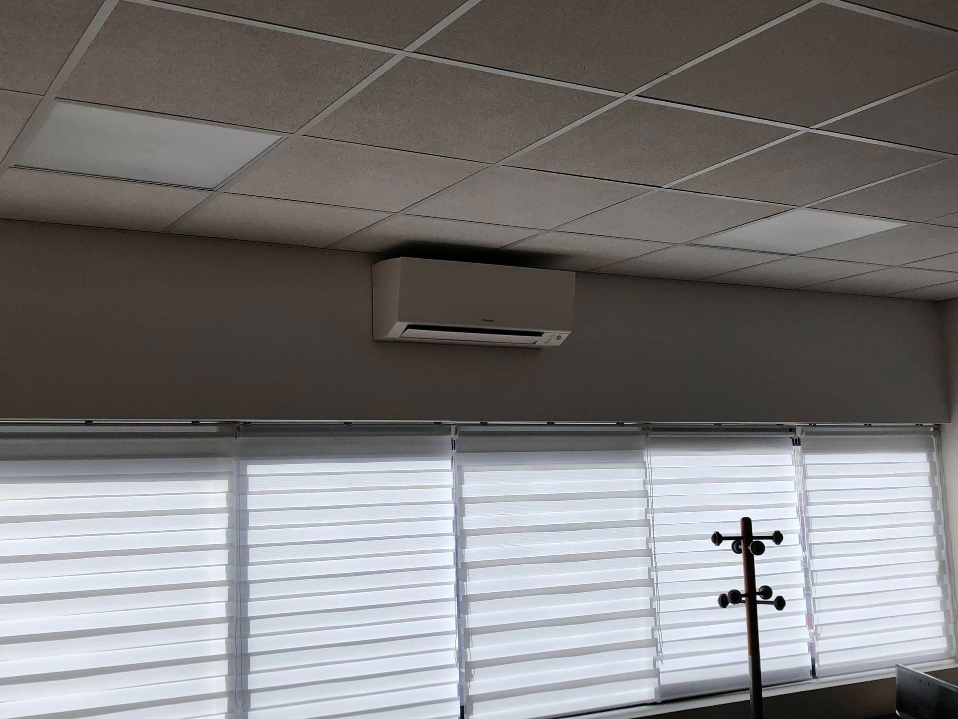 Un climatiseur est fixé au mur, au-dessus de stores. Un porte-manteau est placé devant la fenêtre.