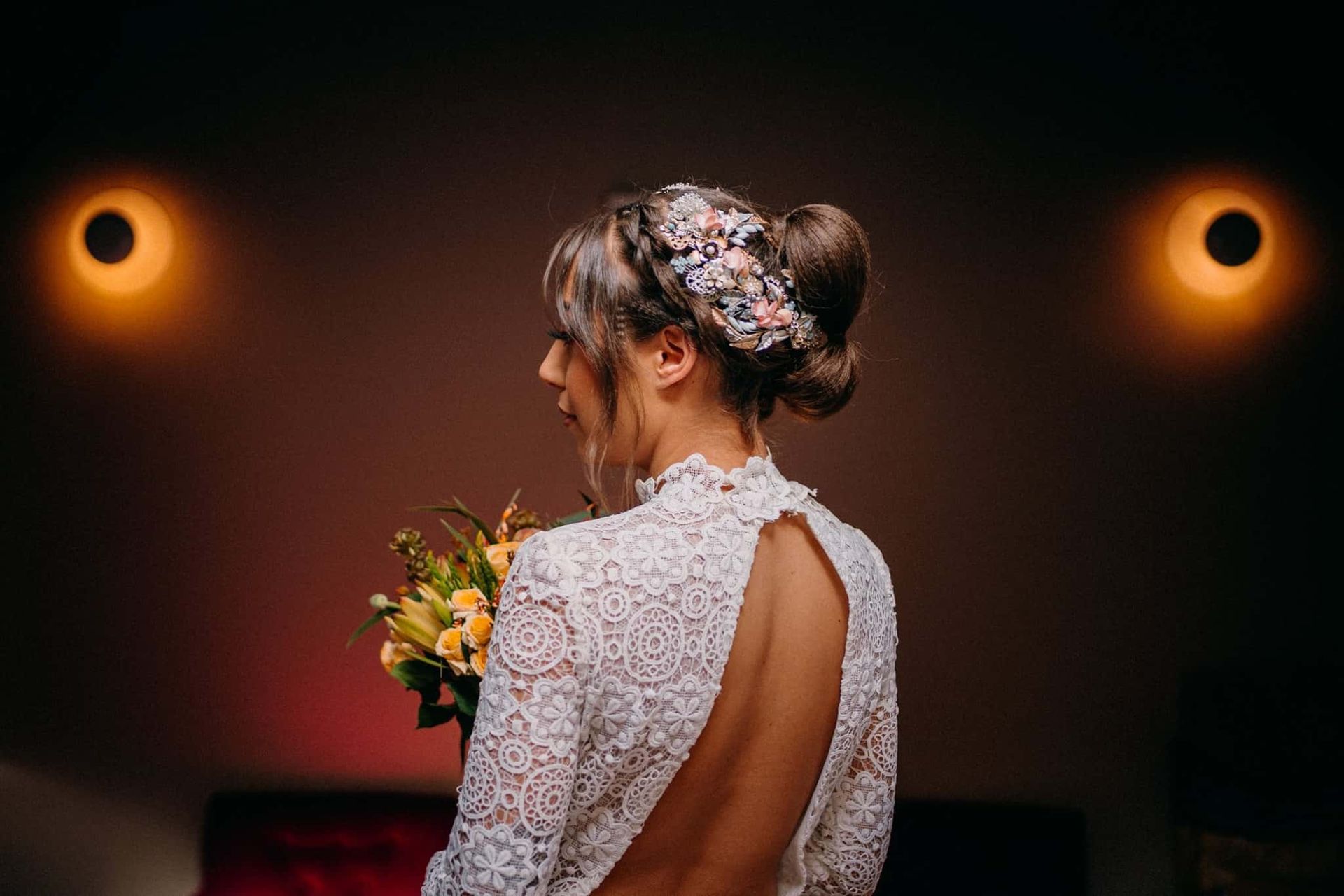Novia con vestido de encaje, espalda descubierta, sostiene ramo, recogido en la cabeza con tocado floral, iluminado por apliques de pared.