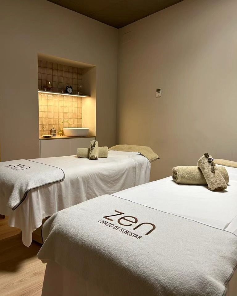 Dos mesas de masaje en una sala de spa con paredes neutrales y un rincón iluminado; las toallas están enrolladas sobre las mesas.