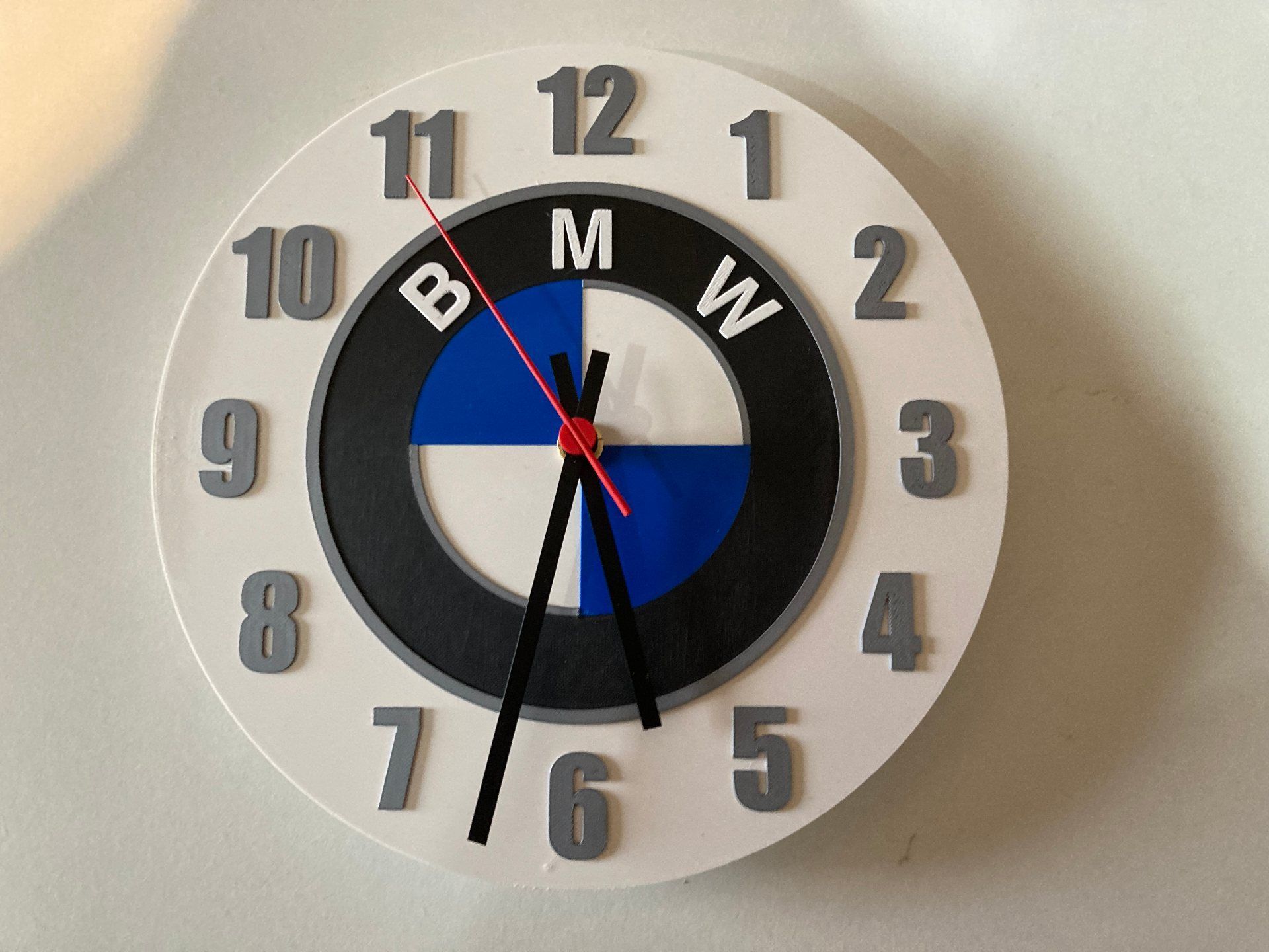 Orologio a muro con logo BMW, quadrante nero, bianco e blu e lancette nere, che segna circa le 6:30.