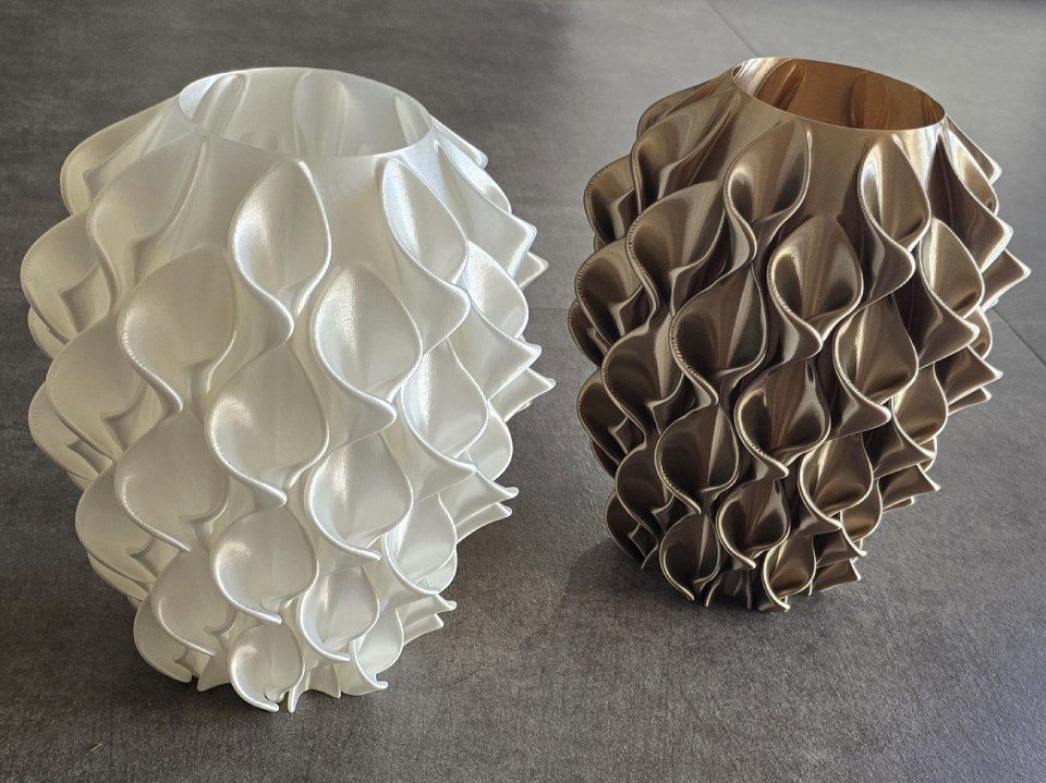 Due vasi decorativi con superficie materica, uno bianco e uno color bronzo, su una superficie grigia.