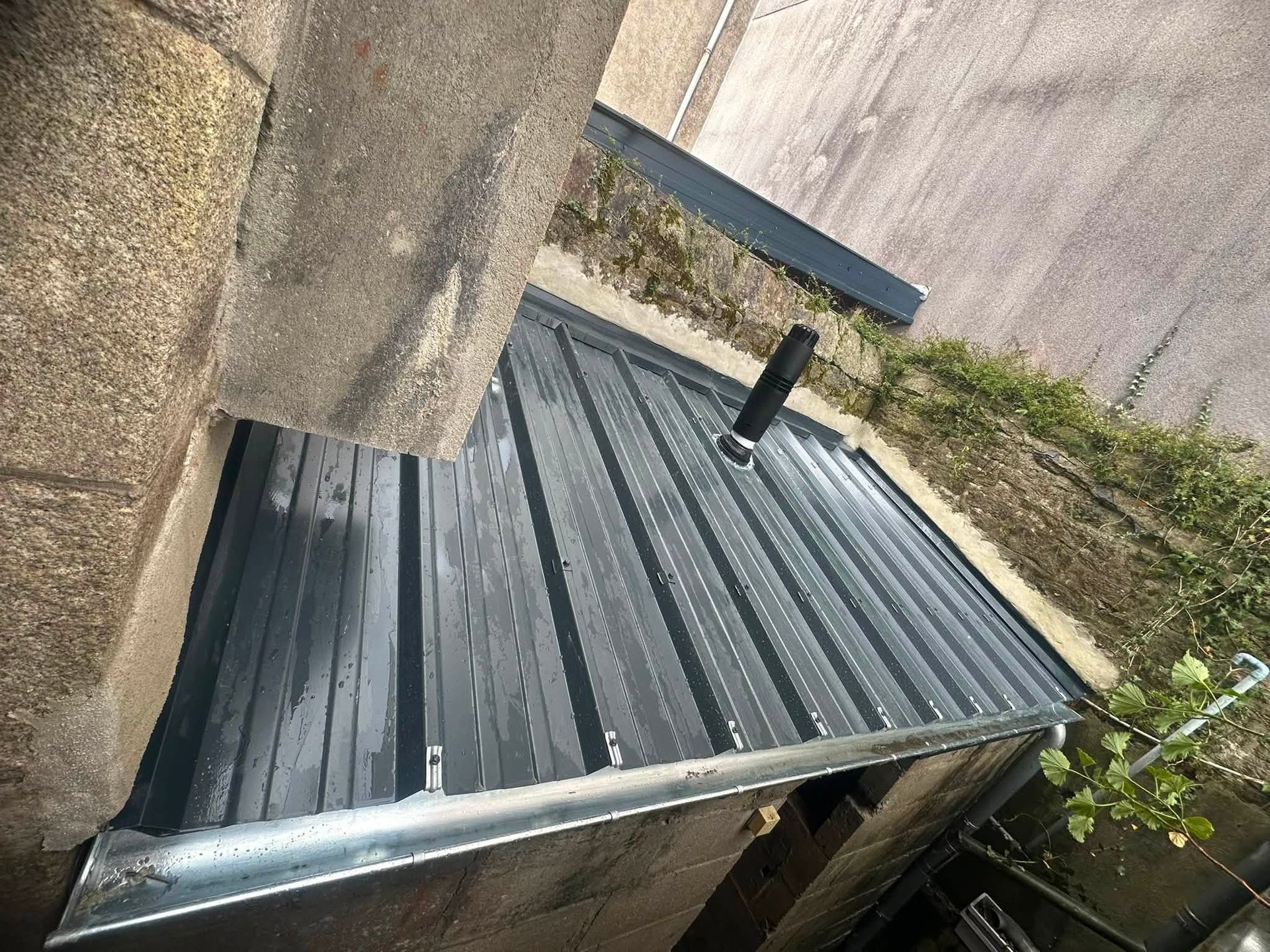 Un toit en tôle ondulée jouxtant un mur en pierre, avec un conduit de ventilation et de la végétation à proximité.