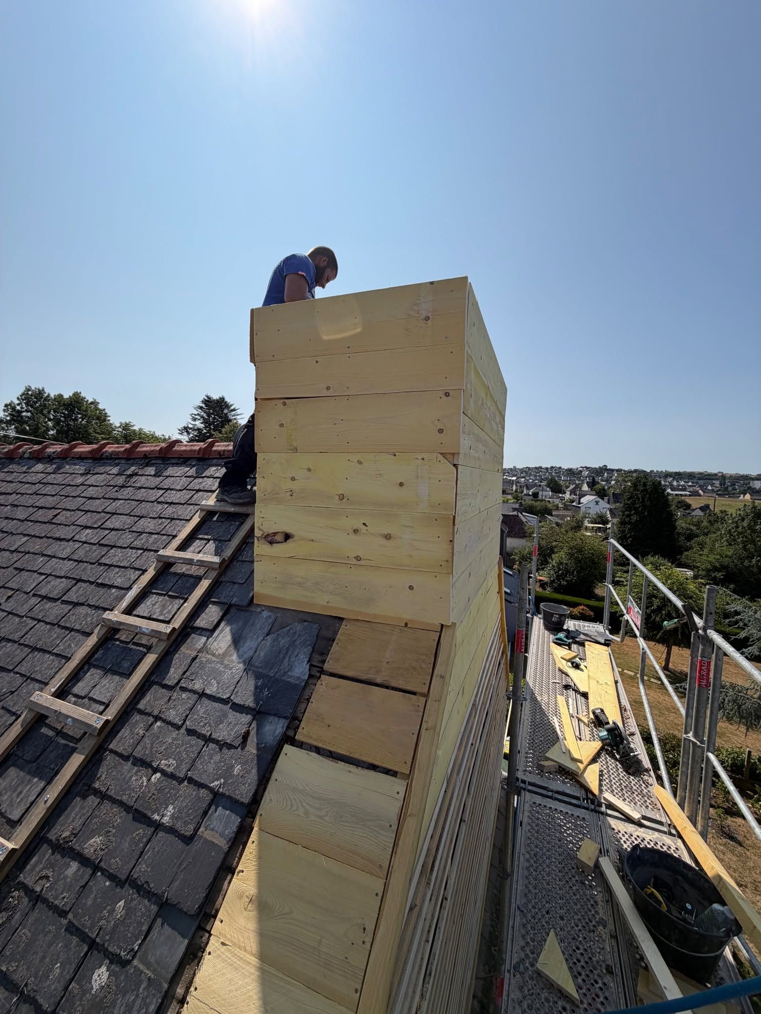 Un ouvrier se trouve au sommet d'une cheminée en construction sur un toit, une échelle appuyée contre celui-ci.