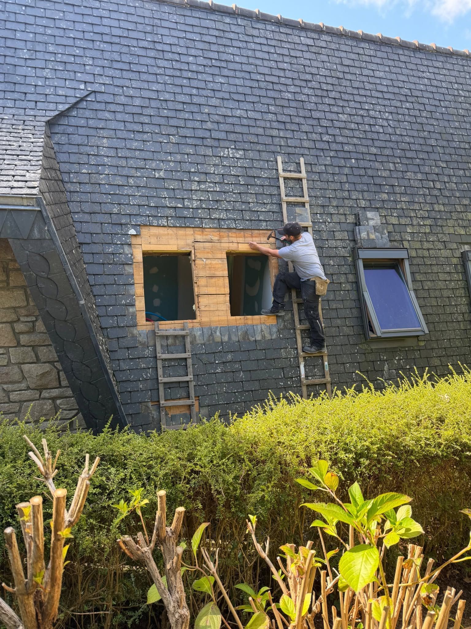 Une personne sur une échelle peint les encadrements de fenêtres d'une maison au toit d'ardoise.