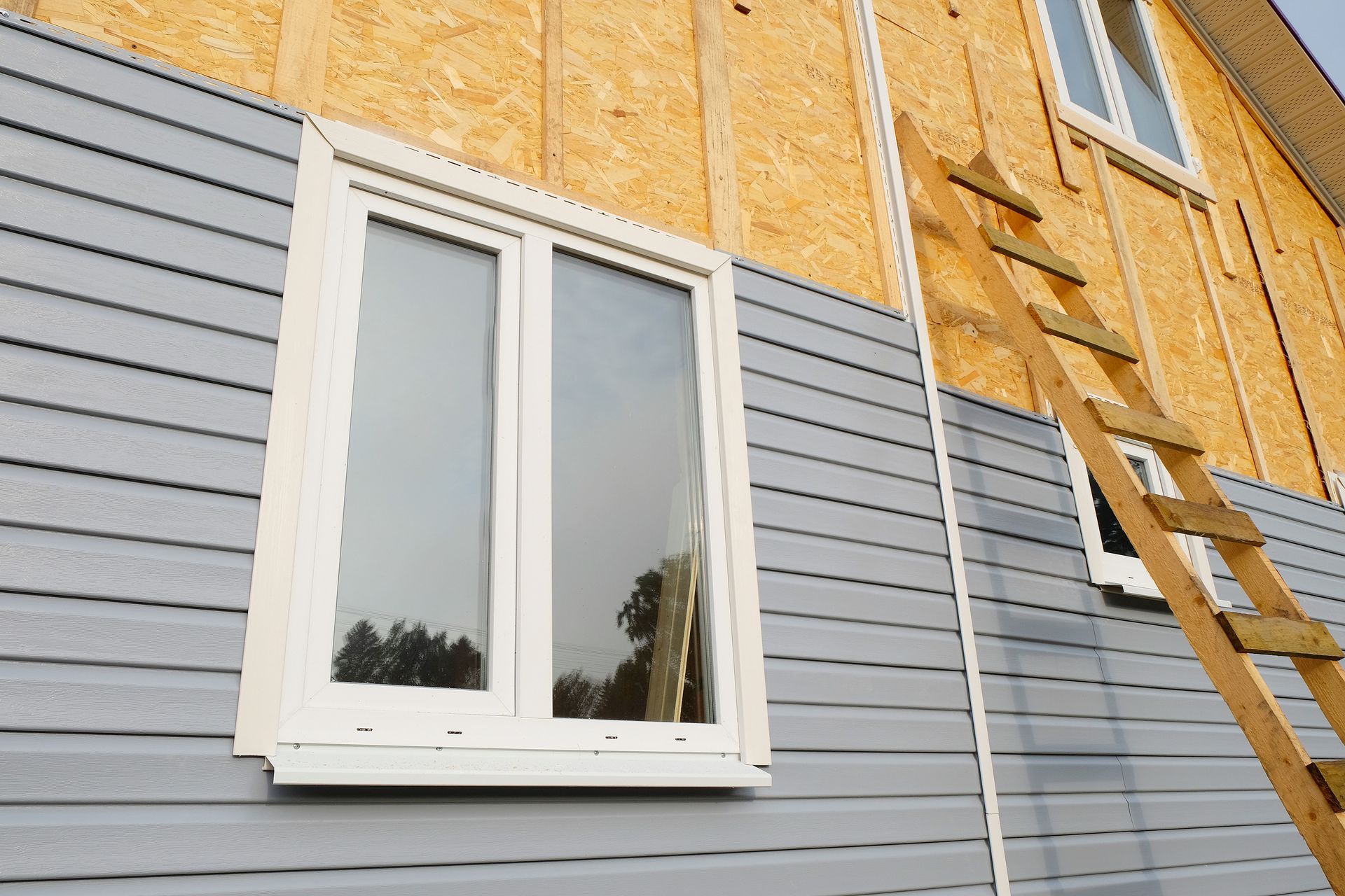 Fenêtre sur du bardage gris à côté d'un revêtement en bois apparent et d'une échelle sur la façade d'une maison.