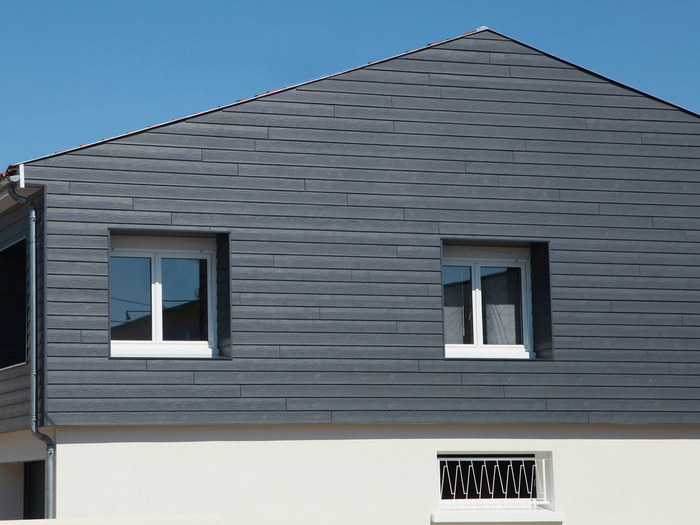 Façade de maison grise avec deux fenêtres sous un toit en bardeaux sombres.