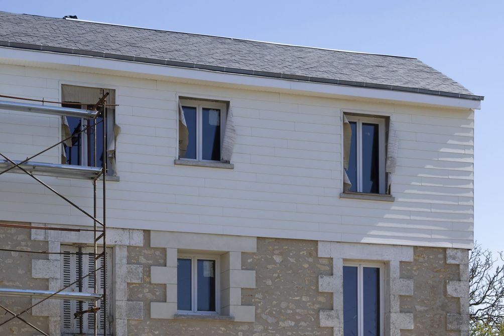 Maison à deux étages avec un bardage supérieur blanc, une façade inférieure en pierre et des échafaudages sur le côté.