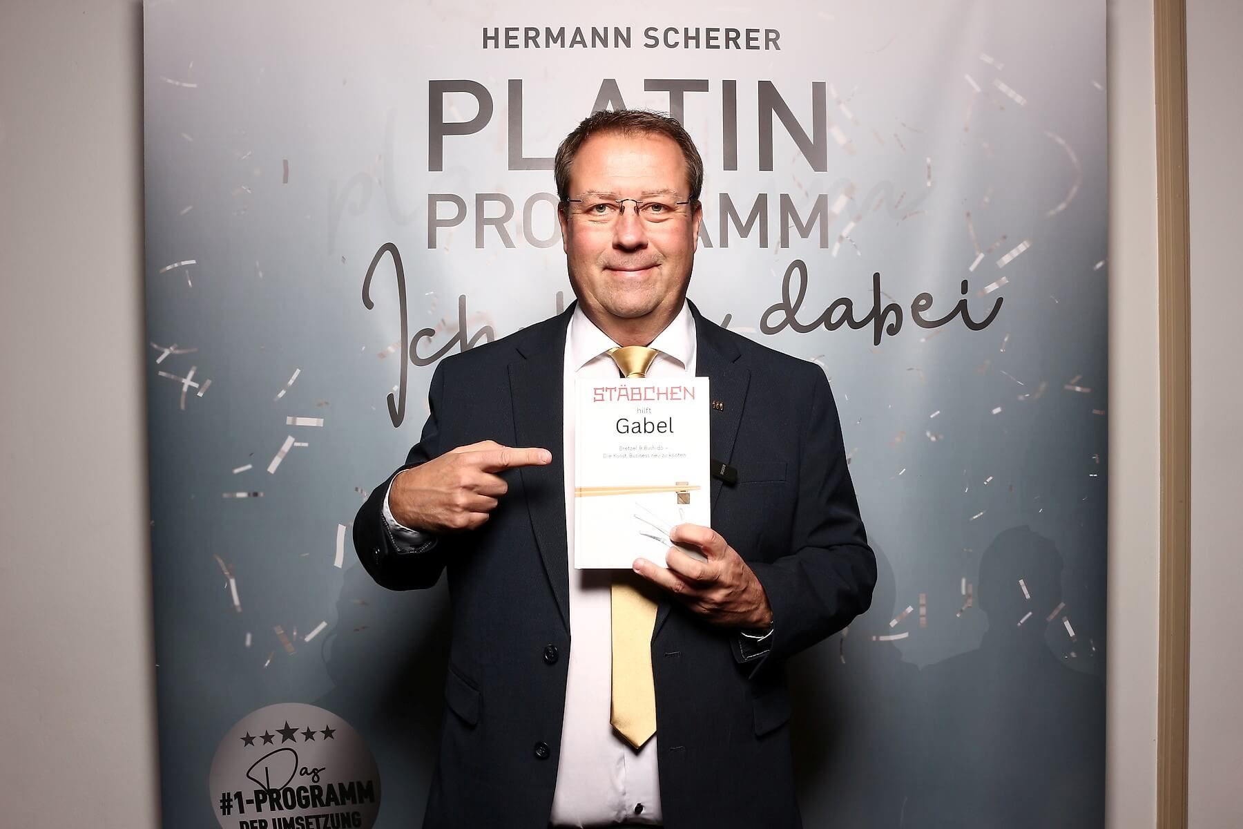 Birger Nispel im Anzug hält ein Buch und zeigt darauf. Hinter ihm hängt ein Banner mit der Aufschrift „Platin Programm“.
